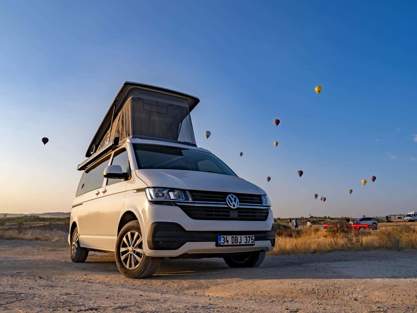 Cappadocia auf weißem VW T6.1 bei Sonnenaufgang in der kappadokischen Ebene, Aufstelldach geöffnet, Heißluftballons am Himmel