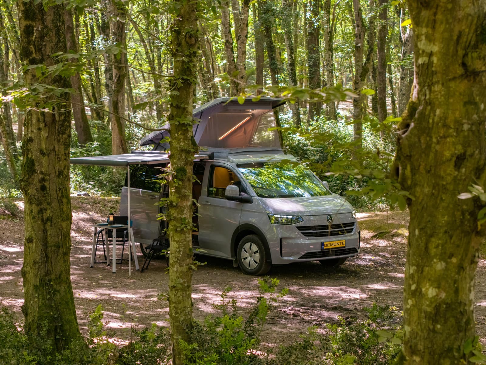 New Cappadocia auf dem VW Transporter der neuen Generation im Wald, Aufstelldach und Markise geöffnet, Campingtisch darunter
