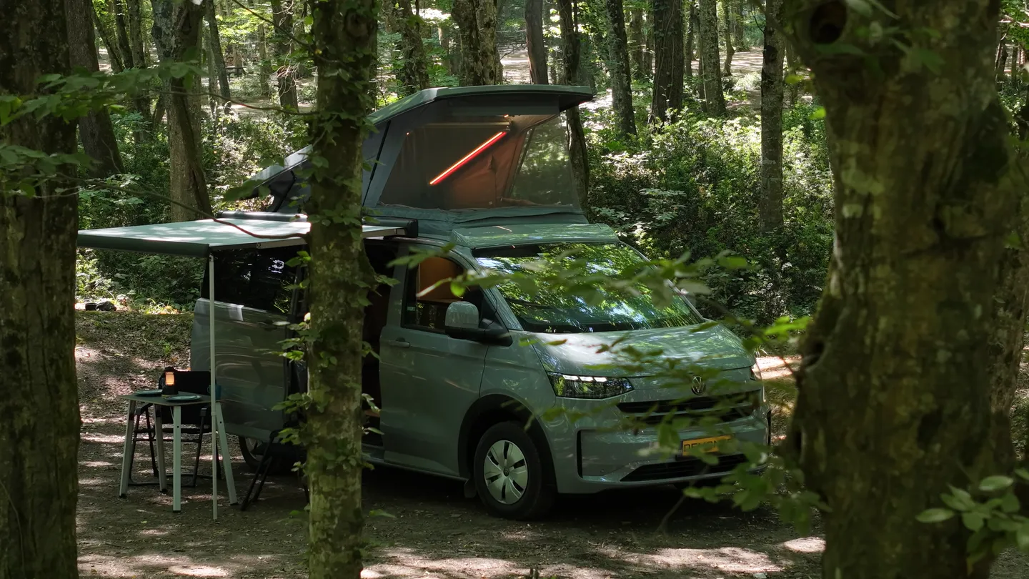 Die Kamera gleitet zwischen den Bäumen hindurch und enthüllt einen Demonte-Camper mit aufgestelltem Dach und Markise